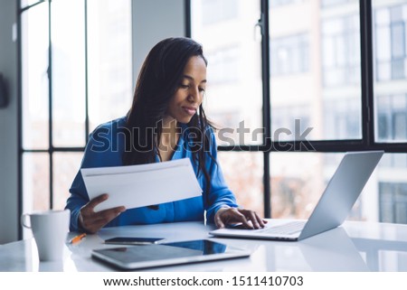 Concentrated brunette African American businesswoman with white envelop in hand sitting at round table and typing on laptop in cafe