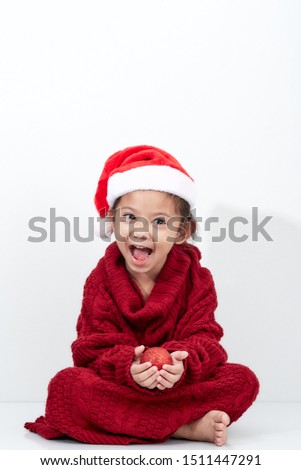 Cute little asian girl holding a red christmas ball in christmas knitted sweater and santa hat studio shot on white background