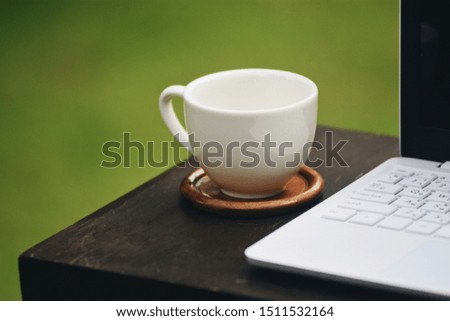 The picture of white laptop, a coffee cup above wooden table on blurred green grass background. Selective focus