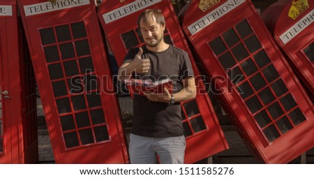 Smiling man showing thumbs up, standing against British telephone, studying English.