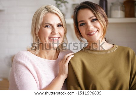 Family portrait of charming young woman posing indoors with her cheerful forty year old mother. Attractive blonde female smiling broadly, keeping hand on shoulder of her cute teenage daughter
