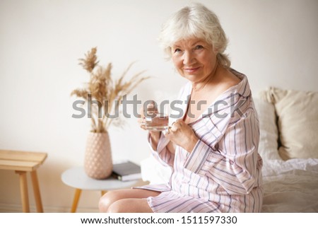 Sleepy happy gray haired European woman pensioner in stylish striped night gown sitting in bedroom on bed, looking at camera, drinking fresh water from glass. Healthy habits, age and retirement