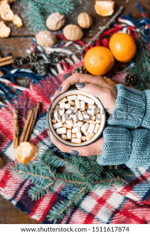 Overview of female hands holding mug of hot drink with marshmallows over warm checkered scarf
