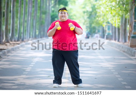 Picture of young fat man exercising with dumbbells while standing on the road 