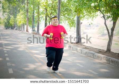 Picture of young fat man holding two dumbbells while jogging on the road