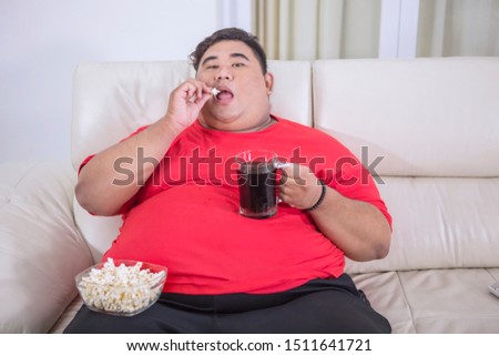 Picture of overweight man watching TV while eating popcorn and drinking cold cola on the couch 