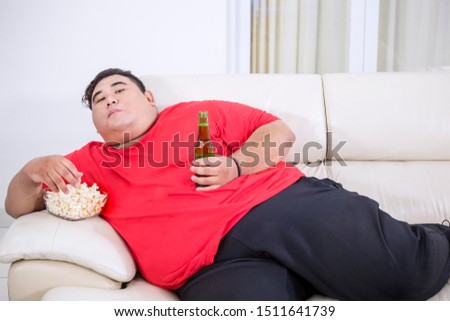 Picture of overweight man watching TV while eating popcorn and drinking beer on the couch 