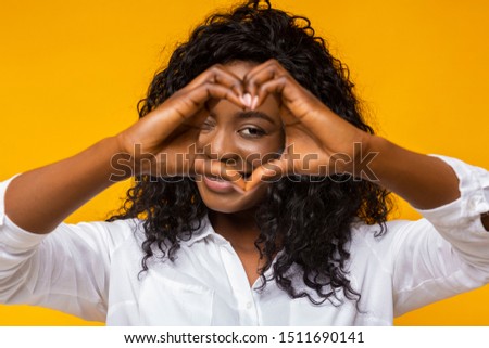 Smiling loving black woman shaping hands like heart over her face, yellow background