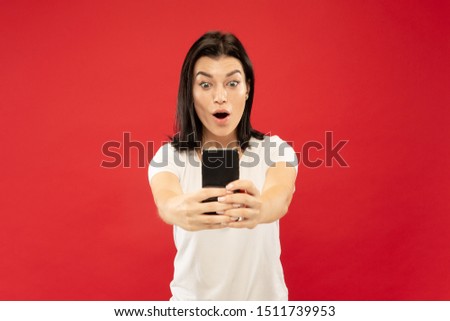 Caucasian young woman's half-length portrait on red studio background. Beautiful female model in white shirt. Concept of human emotions, facial expression. Making selfie or her own vlog, blog.