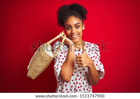 African american woman wearing fashion dress holding wicker bag over isolated red background happy with big smile doing ok sign, thumb up with fingers, excellent sign