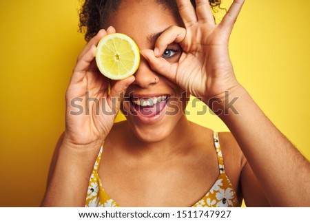 Young african american woman holding slice of lemon over isolated yellow background with happy face smiling doing ok sign with hand on eye looking through fingers