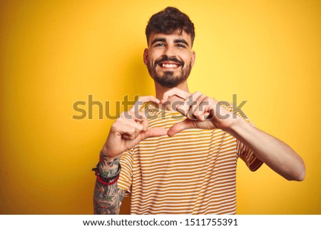 Young man with tattoo wearing striped t-shirt standing over isolated yellow background smiling in love doing heart symbol shape with hands. Romantic concept.