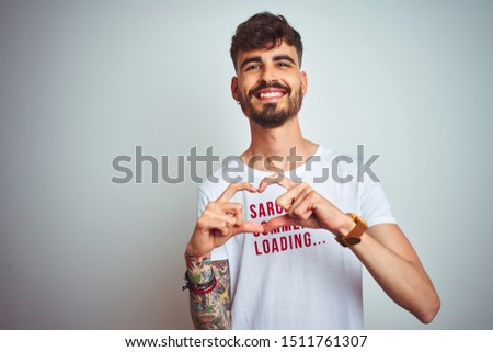 Young man with tattoo wearing fanny t-shirt standing over isolated white background smiling in love showing heart symbol and shape with hands. Romantic concept.