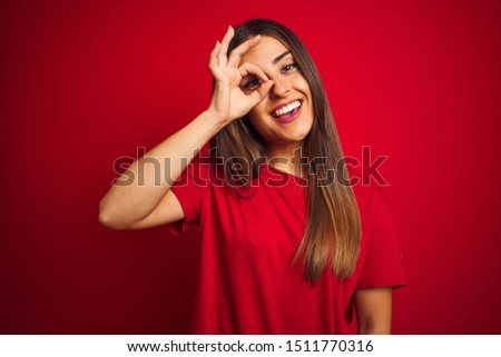 Young beautiful woman wearing t-shirt standing over isolated red background doing ok gesture with hand smiling, eye looking through fingers with happy face.