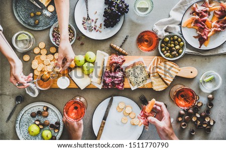 Mid-summer picnic with wine and snacks. Flat-lay of charcuterie and cheese board, rose wine, nuts, olives and peoples hands with snacks over concrete table background, top view