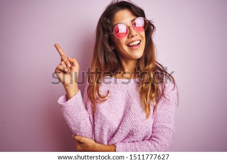 Young beautiful woman wearing fashion sunglasses standing over pink isolated background with a big smile on face, pointing with hand and finger to the side looking at the camera.
