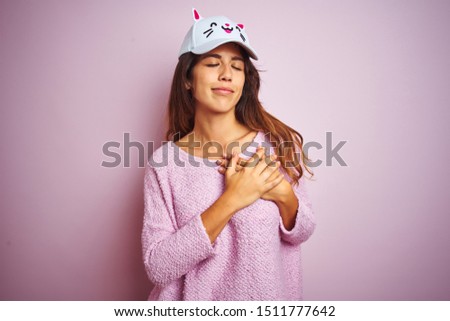 Young beautiful woman wearing funny cat cap standing over pink isolated background smiling with hands on chest with closed eyes and grateful gesture on face. Health concept.