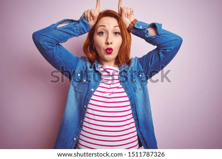 Beautiful redhead woman wearing denim shirt and striped t-shirt over isolated pink background doing funny gesture with finger over head as bull horns