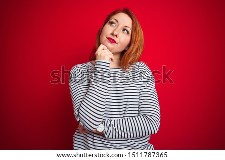 Young redhead woman wearing strapes navy shirt standing over red isolated background with hand on chin thinking about question, pensive expression. Smiling with thoughtful face. Doubt concept.