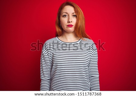Young redhead woman wearing strapes navy shirt standing over red isolated background Relaxed with serious expression on face. Simple and natural looking at the camera.