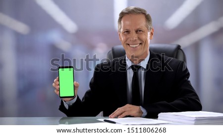 Joyful male boss in suit showing green screen smartphone at camera, business app