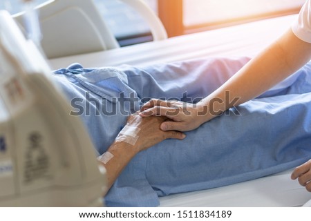 Caregiver holding elderly senior patient (ageing old adult person) hand lying in hospital bed or nursing hospice, geriatrician palliative home, while caretaker having  medical health care service   Royalty-Free Stock Photo #1511834189