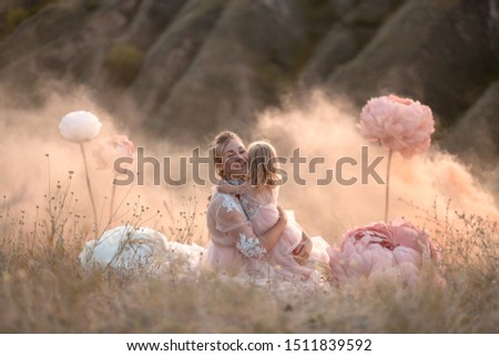 A little girl hugs mom sitting in a field surrounded by unreal big pink decorative flowers.