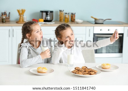 Portrait of cute twin girls with milk taking selfie in kitchen