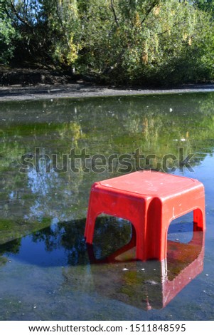 Small red plastic stool in the middle of a beautiful pond landscape view, abstract play objects and nature.
Land Art Photography.