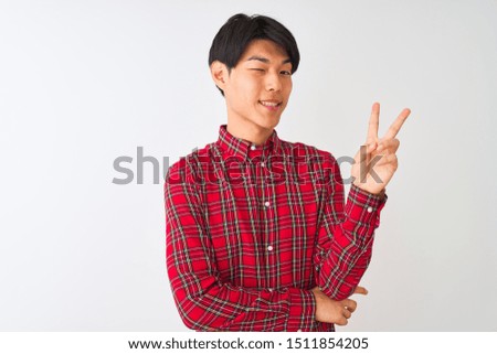 Young chinese man wearing casual red shirt standing over isolated white background smiling with happy face winking at the camera doing victory sign. Number two.