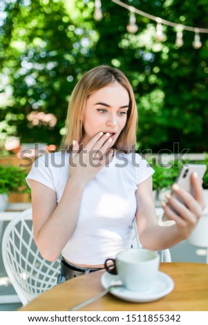 Picture of young shocked excited woman sitting in cafe outdors in park drinking wine using mobile phone.