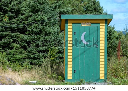 A small yellow and green wooden building with a sign that spells out outhouse over the green door. The outdoor toilet has a moon shape on the exterior side of the door. The park bonnet house is old.