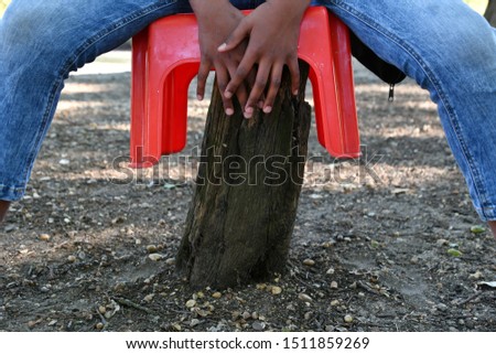 Kid seating on a small red plastic stool on top of a small broken tree trunk, abstract play objects, people and nature.
Land Art Photography.