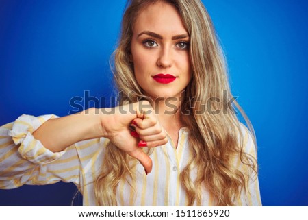 Young beautiful woman wearing striped shirt standing over blue isolated background with angry face, negative sign showing dislike with thumbs down, rejection concept