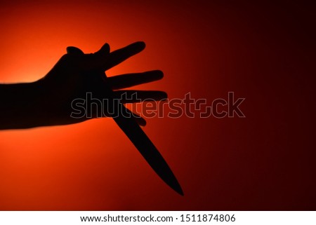 The left hand of a person holding a sharp knife on a red background and lit by a light bulb in a dark atmosphere