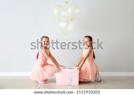 Portrait of cute twin girls with air balloons and big gift near light wall
