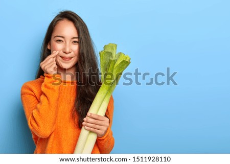 Photo of lovely Asian woman makes korean like hand sign, holds green leek bought in grocers shop, wears orange sweater, going to cook vegetarian dish, poses over blue wall with blank copy space