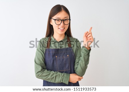 Young chinese shopkeeper woman wearing apron and glasses over isolated white background with a big smile on face, pointing with hand and finger to the side looking at the camera.