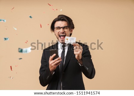 Portrait of a handsome excited young businessman wearing suit standing isolated over beige background, celebrating while showing plastic credit card and using mobile phone