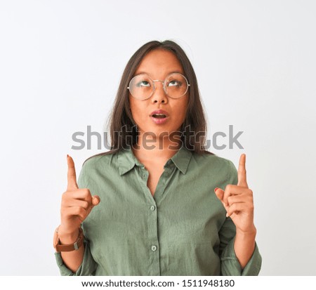 Young chinese woman wearing green shirt and glasses over isolated white background amazed and surprised looking up and pointing with fingers and raised arms.