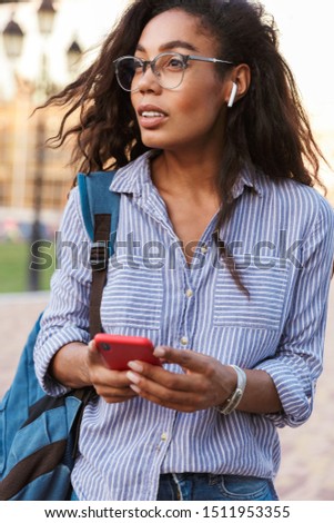 Attractive young african woman student carrying backpack walking outdoors, listening to music with wireless earphones while using mobile phone