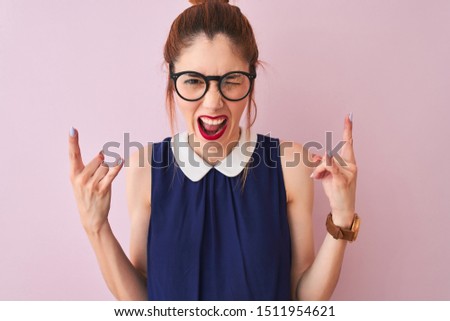 Redhead woman with pigtail wearing elegant dress and glasses over isolated pink background shouting with crazy expression doing rock symbol with hands up. Music star. Heavy concept.