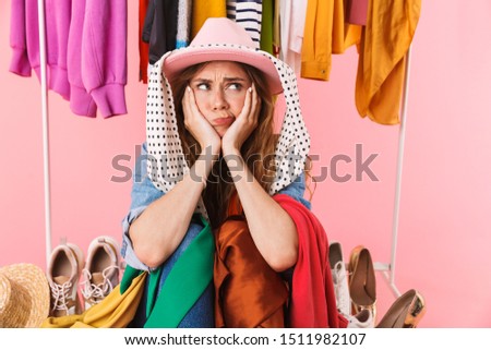 Photo of puzzled young woman wearing hat sitting near bunch of clothes and shoes isolated over pink background