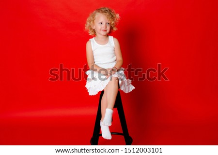 Modest little blonde girl in white dress sitting on chair isolated on red