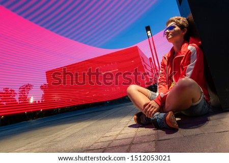 Photo of young woman with short haircut, in sunglasses, sits on floor, urban landscape on background. Drawing with light. Long exposure. Artistic abstraction. Freezelight