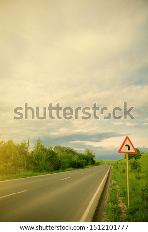
Road sign indicating left turn, blue sky with clouds and green trees 