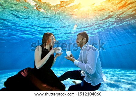 An unusual loving couple, a guy and a girl, look at each other, sitting underwater at the bottom of the pool with white cups in their hands. Surrealism. The concept of tranquility and serenity Royalty-Free Stock Photo #1512104060