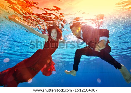 Unusual underwater wedding. A beautiful redhead girl in a red dress and a mixed race guy in a suit are swimming and playing underwater in the pool. Fashion portrait.