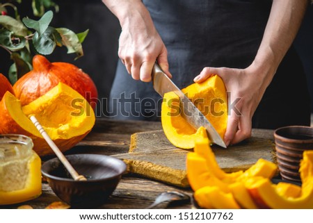 A woman cook cuts an orange pumpkin with a knife into slices for baking on a wooden table. Concept autumn food in a cozy dark kitchen with yellow flowers. Close up