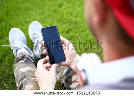 Focus on stylish sensory smartphone in male hands. Guy in casual clothes sitting on green grass and chatting with friends via special mobile app. Empty copy space on gadget screen